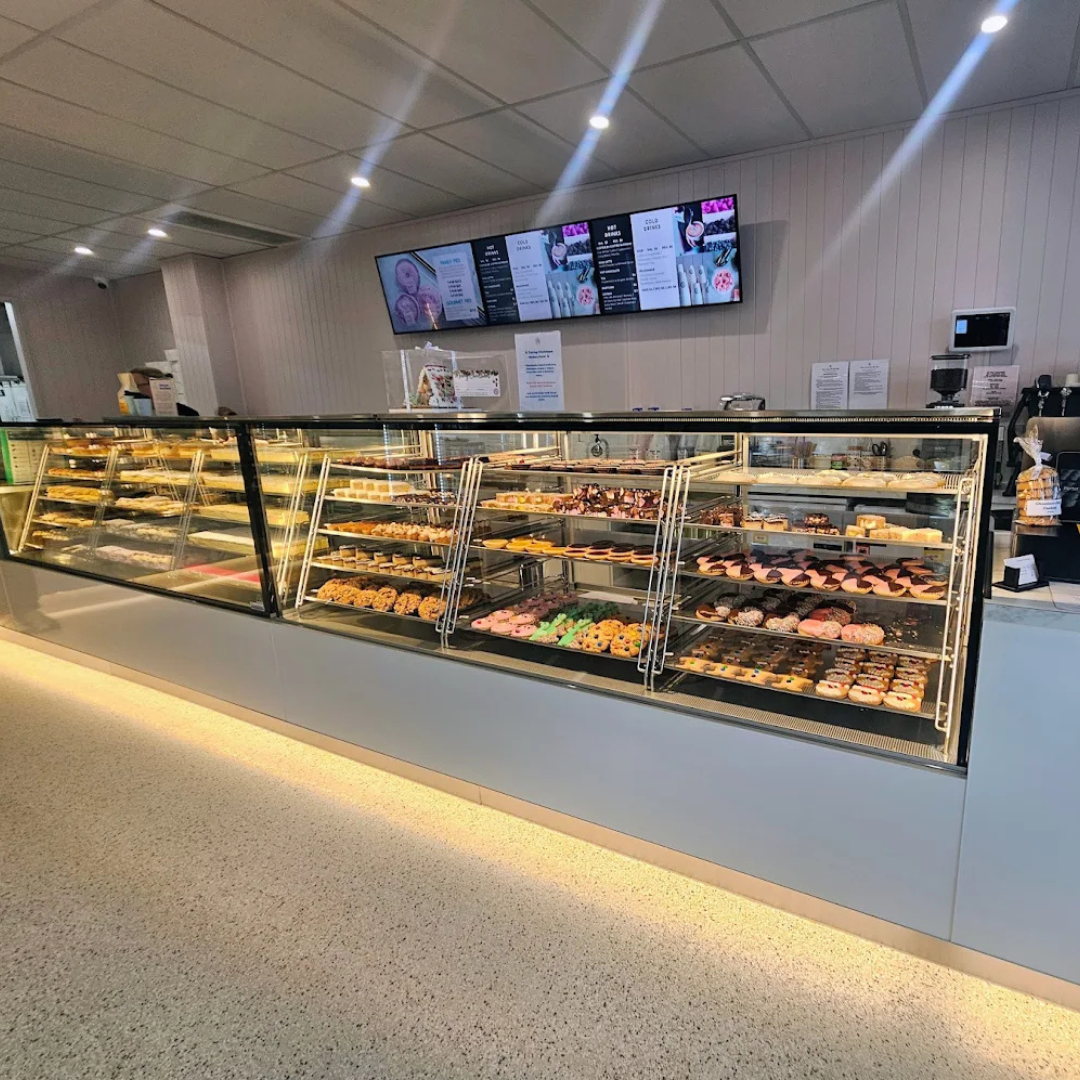A bakery display case filled with assorted pastries, donuts, and cakes in a commercial cafe interior, with digital menu boards on the wall, under cabinet lighting and a tiled floor.