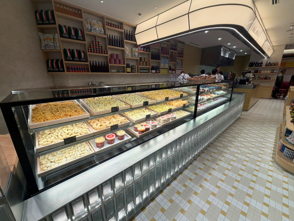A long glass-front bakery display case filled with chilled products, set inside a bright restaurant with tiled floors, shelving, and staff visible in the background.