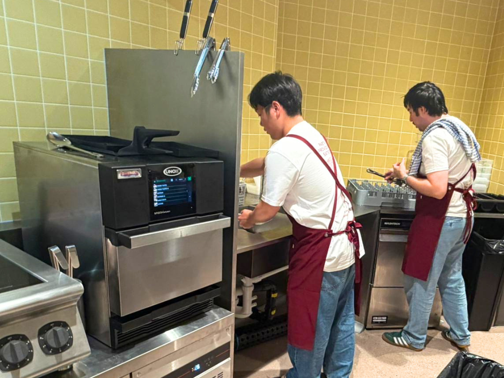 Two people in white shirts and red aprons working in a commercial kitchen, with stainless steel fryers, commercial dishwasher and ovens and yellow tiled walls in the background.