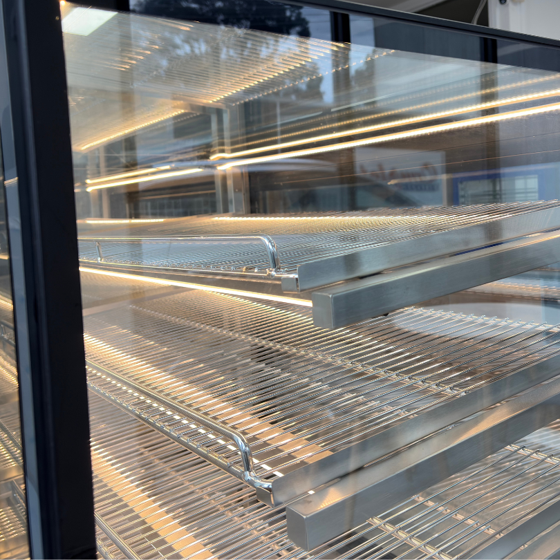 A close-up view of stainless steel wire cooling shelves inside a glass-door refrigerator or display case, with warm interior lighting reflecting off the metal and glass.