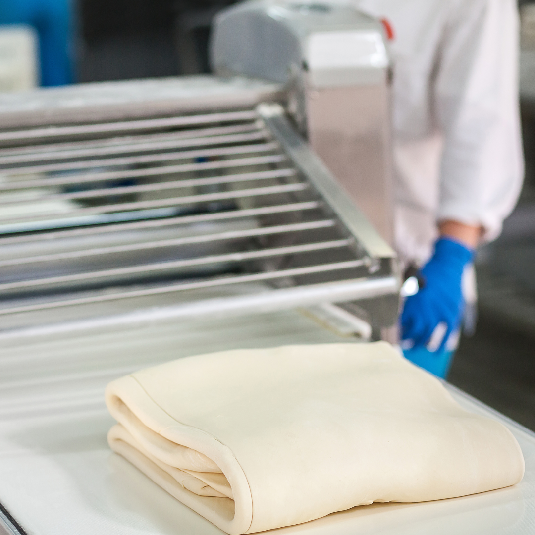 close up of dough about to move through bakery pastry sheeter, featuring long conveyor belts and  rollers on a floor-standing base